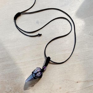 May include: A handmade necklace featuring a pointed, light purple crystal pendant wrapped in black string. The necklace has a black cord and a small purple bead near the pendant. The background is a light wood surface.