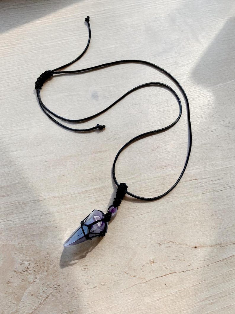 May include: A handmade necklace featuring a pointed, light purple crystal pendant wrapped in black string. The necklace has a black cord and a small purple bead near the pendant. The background is a light wood surface.