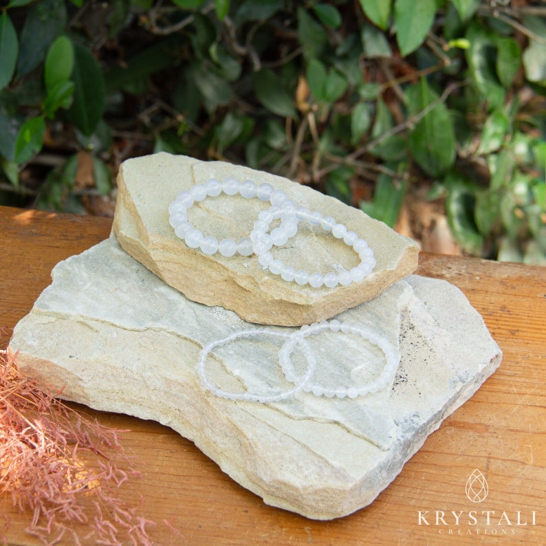 May include: Four white gemstone bracelets stacked on top of two light brown rocks. The bracelets are made of small, round beads.