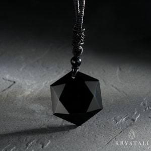 K&ouml;nnte beinhalten: Eine schwarze geometrische Anh&auml;nger Halskette mit facettiertem Design. Der Anh&auml;nger h&auml;ngt an einer schwarzen Kordel mit einem Gleitknoten und einer kleinen schwarzen Perle. Die Halskette wird auf einer strukturierten grauen Oberfl&auml;che pr&auml;sentiert.
