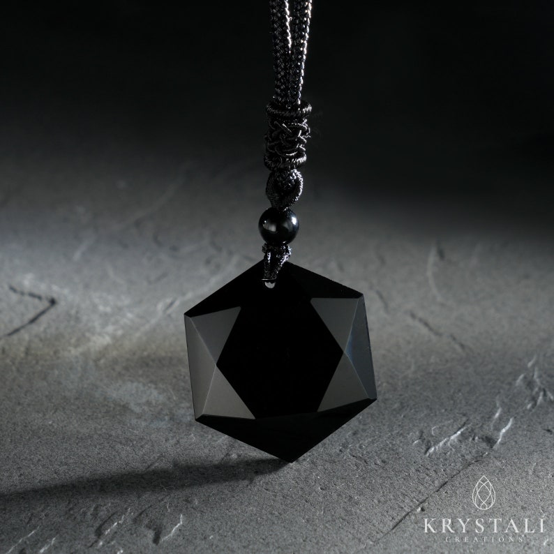 K&ouml;nnte beinhalten: Eine schwarze geometrische Anh&auml;nger Halskette mit facettiertem Design. Der Anh&auml;nger h&auml;ngt an einer schwarzen Kordel mit einem Gleitknoten und einer kleinen schwarzen Perle. Die Halskette wird auf einer strukturierten grauen Oberfl&auml;che pr&auml;sentiert.