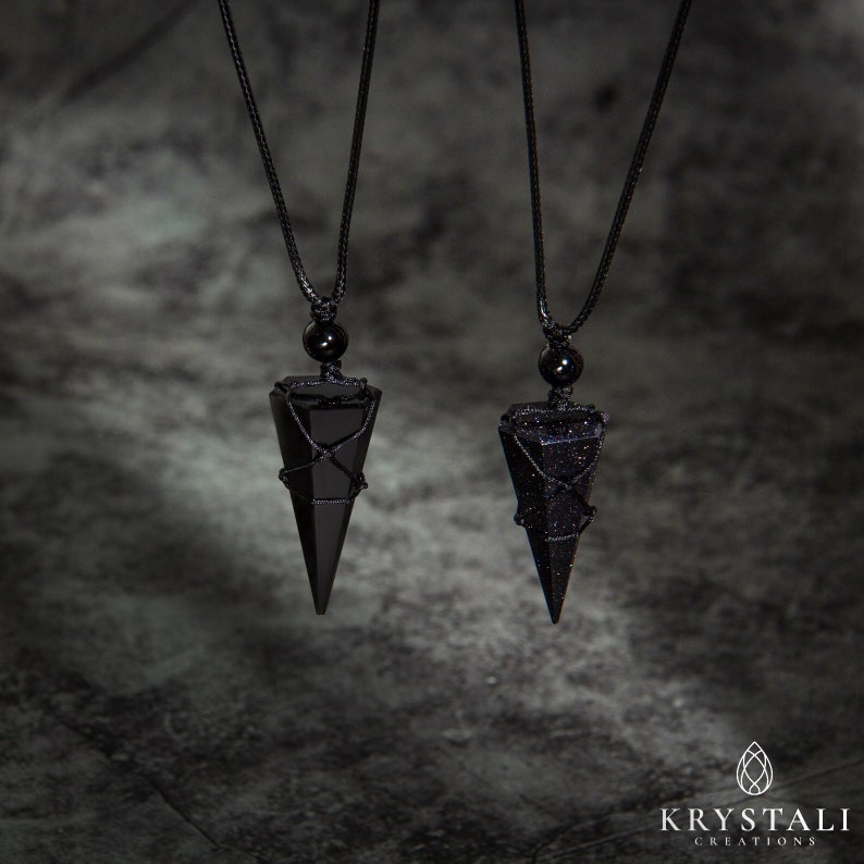 May include: Two black crystal pendant necklaces. Each pendant is a triangular shape, wrapped in black wire, and suspended from a black cord. One pendant is solid black, the other is a dark blue with glittery inclusions. The necklaces are displayed against a dark gray background.