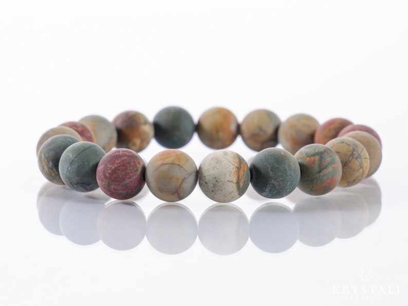 May include: A beaded bracelet featuring large, round beads in shades of green, brown, red, and tan. The beads have a matte finish and are strung together to form a circular shape. The bracelet is displayed on a white surface.