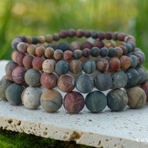 May include: Stacked bracelets featuring natural stone beads in shades of brown, gray, and green. The beads are round and vary in size, creating a layered effect. The bracelets are displayed on a light-colored surface with a blurred green background.