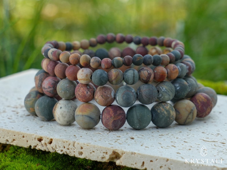May include: Stacked bracelets featuring natural stone beads in shades of brown, gray, and green. The beads are round and vary in size, creating a layered effect. The bracelets are displayed on a light-colored surface with a blurred green background.