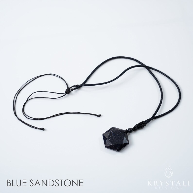 K&ouml;nnte beinhalten: Halskette mit Anh&auml;nger aus blauem Sandstein und schwarzem Band. Der Anh&auml;nger hat eine facettierte, sechseckige Form mit gesprenkelter Optik. Die Halskette wird vor wei&szlig;em Hintergrund pr&auml;sentiert. Der Text "BLUE SANDSTONE" steht unten.