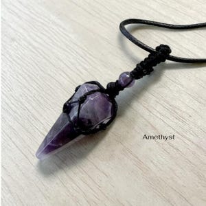 May include: Amethyst crystal pendant with a black cord. The purple crystal is encased in a black woven net. The word "Amethyst" is printed in white on the bottom right.