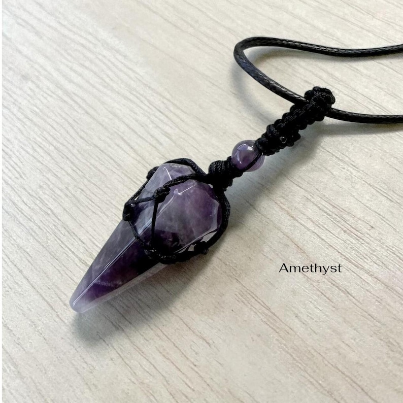 May include: Amethyst crystal pendant with a black cord. The purple crystal is encased in a black woven net. The word "Amethyst" is printed in white on the bottom right.