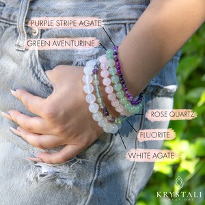 May include: A stack of five beaded bracelets in various colors, including white, green, pink, and purple. The bracelets are labeled with their corresponding gemstone names: White Agate, Fluorite, Rose Quartz, Green Aventurine, and Purple Stripe Agate.