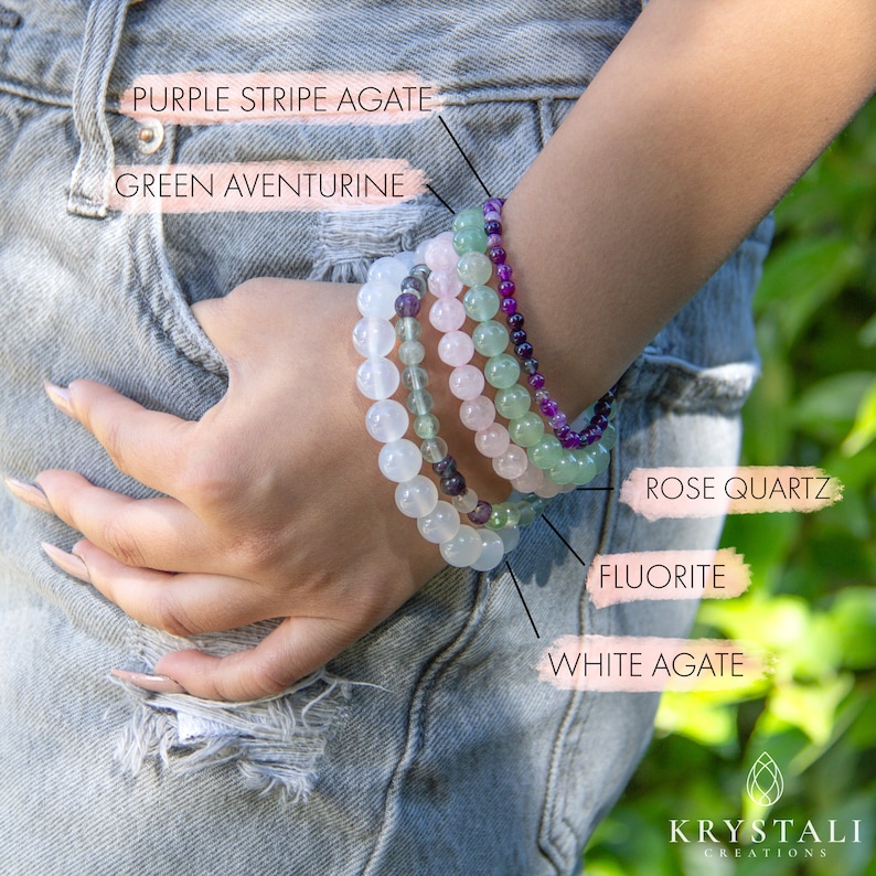 May include: A stack of five beaded bracelets in various colors, including white, green, pink, and purple. The bracelets are labeled with their corresponding gemstone names: White Agate, Fluorite, Rose Quartz, Green Aventurine, and Purple Stripe Agate.