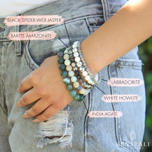 May include: A stack of six bracelets made of various gemstones, including black spider web jasper, matte amazonite, white howlite, labradorite, and India agate.