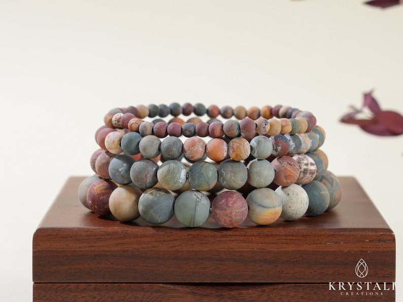 May include: Stacked bracelets with large, round, multi-colored beads. The beads have a natural, earthy appearance with hues of brown, green, and red. The bracelets are displayed on a dark wooden surface.
