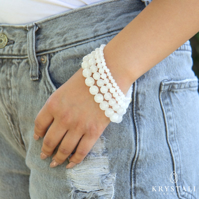 May include: A stack of four white gemstone bracelets with round beads. The bracelets are worn on a person's wrist.