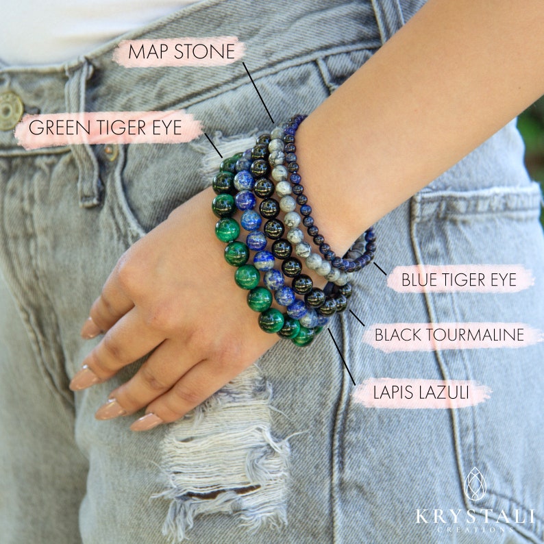 May include: A stack of six beaded bracelets on a person's wrist. The bracelets are made of green, blue, black, and gray stones. The stones are labeled as Green Tiger Eye, Blue Tiger Eye, Black Tourmaline, Lapis Lazuli, and Map Stone.