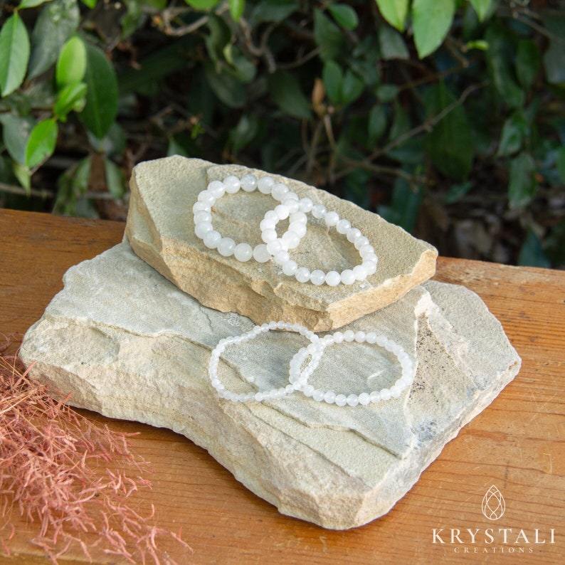 May include: Four white gemstone bracelets stacked on top of two light brown rocks. The bracelets are made of small, round beads.
