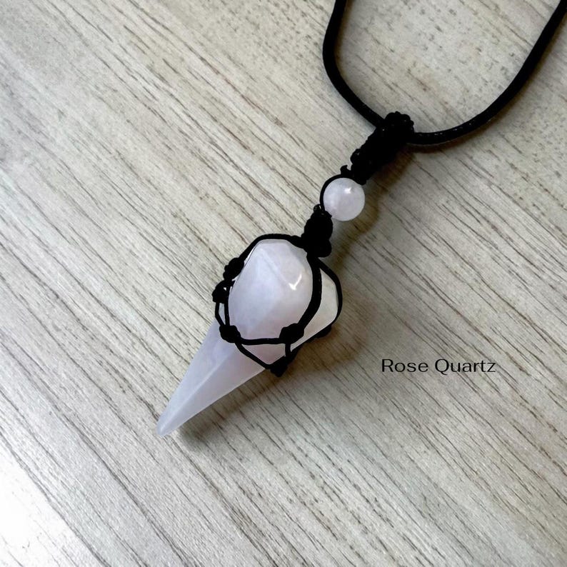 May include: A rose quartz pendulum necklace with a black cord. The light pink crystal is encased in a black woven net. The text "Rose Quartz" is at the bottom right.