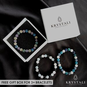 May include: Three beaded bracelets in shades of blue, green, white, and purple are displayed on a black surface. One bracelet is in a white gift box. The box is labeled "Krystali Creations". The text "FREE GIFT BOX FOR 3+ BRACELETS" is at the bottom.