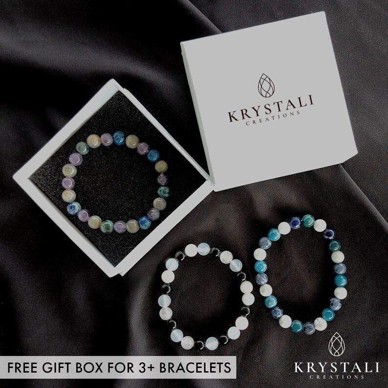 May include: Three beaded bracelets in shades of blue, green, white, and purple are displayed on a black surface. One bracelet is in a white gift box. The box is labeled "Krystali Creations". The text "FREE GIFT BOX FOR 3+ BRACELETS" is at the bottom.