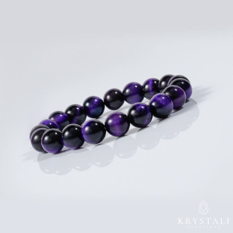 May include: A beaded bracelet featuring polished, round beads in shades of deep purple and black. The bracelet is displayed on a white surface, with the beads reflecting light. The jewelry is a stylish accessory.