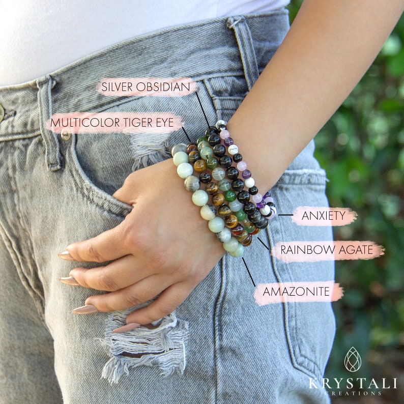 May include: A stack of six beaded bracelets on a person's wrist. The bracelets are made of various gemstones, including amethyst, tiger's eye, amazonite, and rainbow agate. The bracelets are labeled with their corresponding gemstone names.