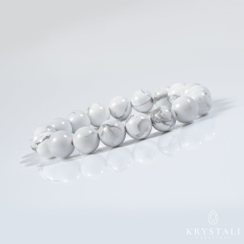 May include: White Howlite beaded bracelet with round beads. The bracelet is laid out on a white surface.
