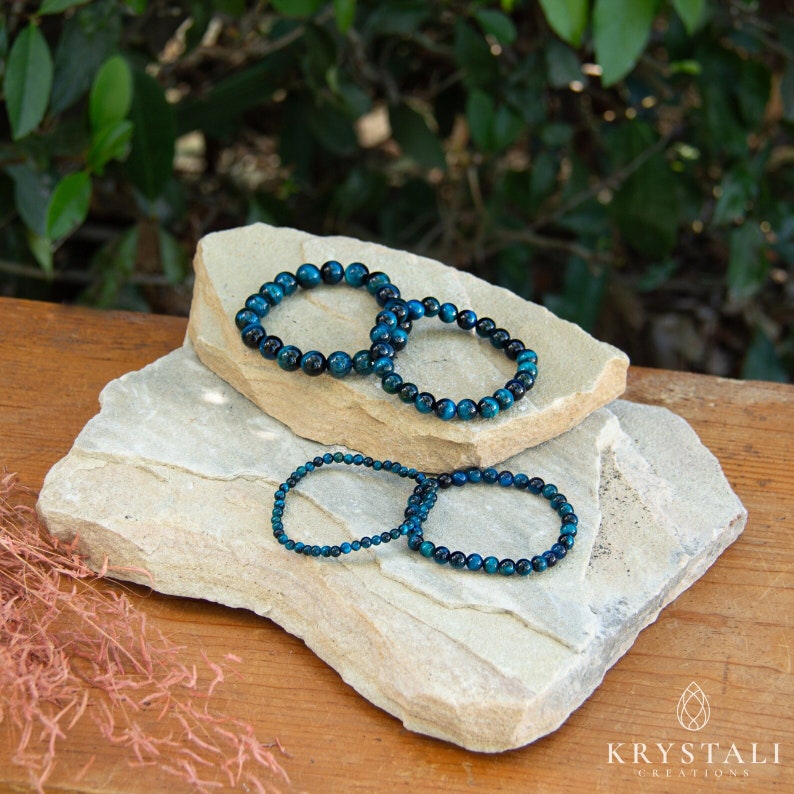 May include: Three blue tiger's eye beaded bracelets stacked on top of each other on a light brown wooden surface. The bracelets are made of small, round beads and are arranged in a circle.