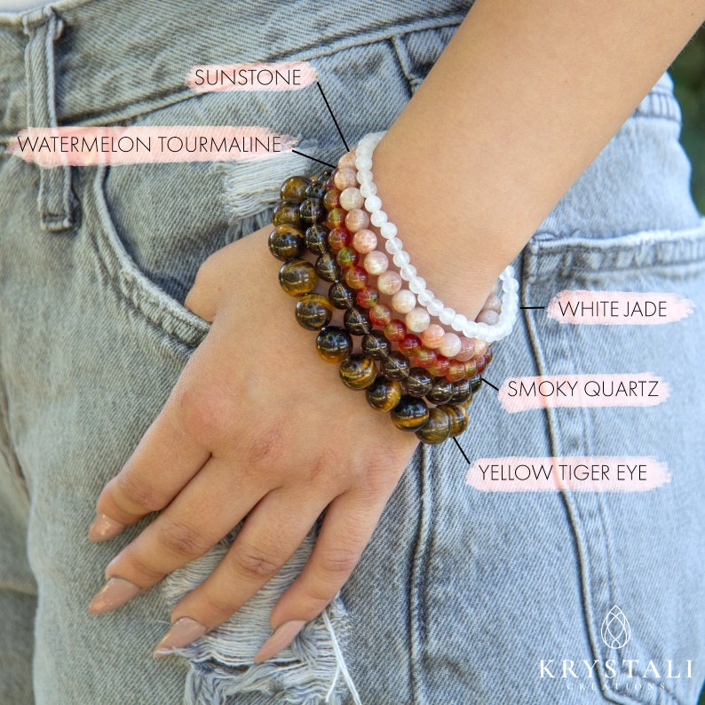 May include: A stack of seven bracelets made of various gemstones, including yellow tiger eye, smoky quartz, white jade, sunstone, and watermelon tourmaline. The bracelets are worn on a person's wrist.