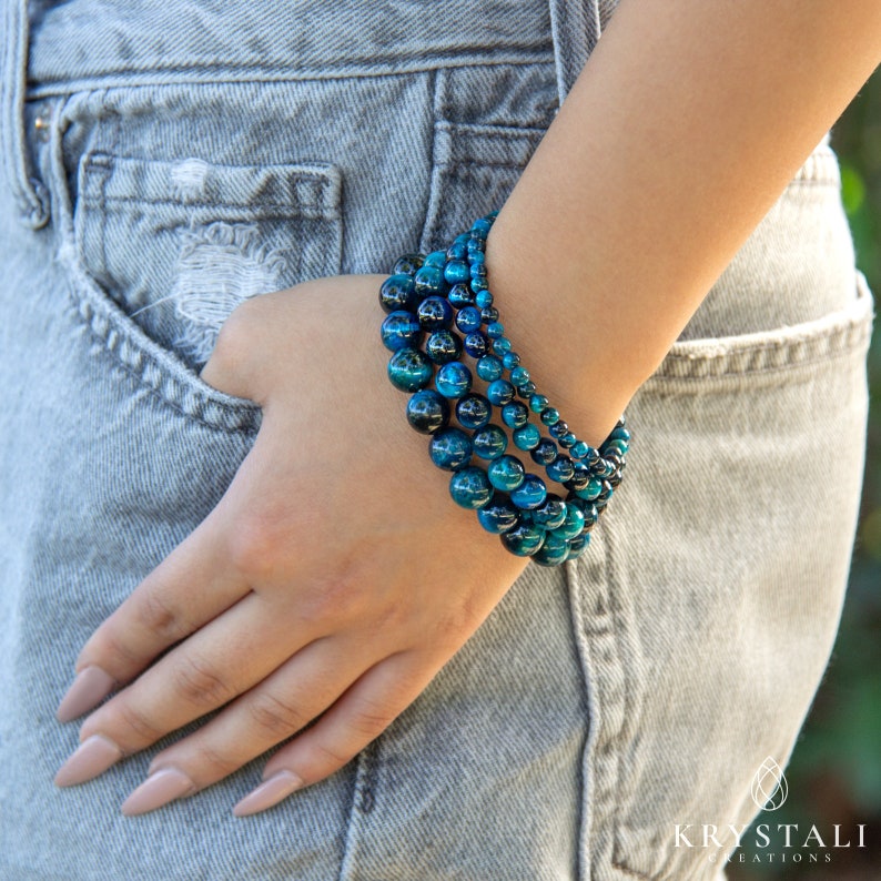 May include: A stack of blue tiger's eye beaded bracelets. The bracelets are layered on a person's wrist.