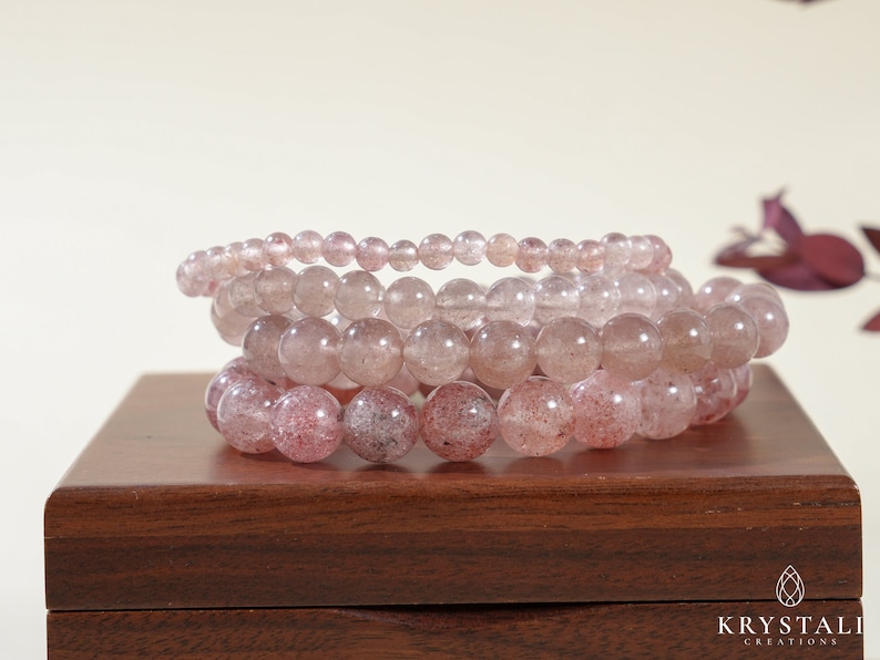 Op de afbeelding: Vier gestapelde armbanden gemaakt van roze en heldere kwarts kralen. De armbanden liggen op een houten doos met de tekst "KRYSTALLI CREATIONS" op de voorkant.