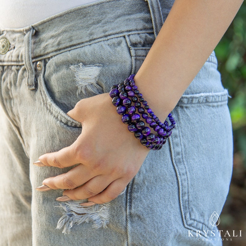 May include: Three beaded bracelets with a mix of purple and black beads are worn on a wrist. The bracelets vary in bead size, creating a layered effect. The person is wearing light blue denim jeans.