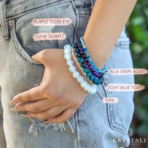 May include: A collection of beaded bracelets featuring various gemstones, including purple tiger eye, clear quartz, blue stripe agate, light blue tiger eye, and opal. The bracelets are stacked on a wrist, showcasing a variety of colors and textures. The brand name "Krystali Creations" is visible.