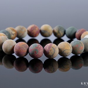 May include: A beaded bracelet featuring round, matte-finish beads in shades of brown, red, green, and cream. The beads have a marbled appearance, creating a natural, earthy aesthetic. The bracelet is displayed on a reflective surface.