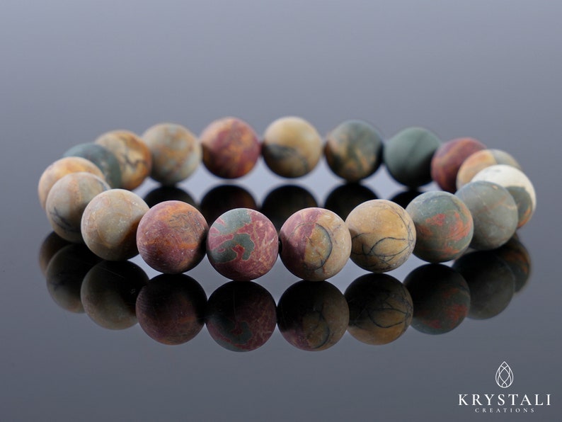May include: A beaded bracelet featuring round, matte-finish beads in shades of brown, red, green, and cream. The beads have a marbled appearance, creating a natural, earthy aesthetic. The bracelet is displayed on a reflective surface.