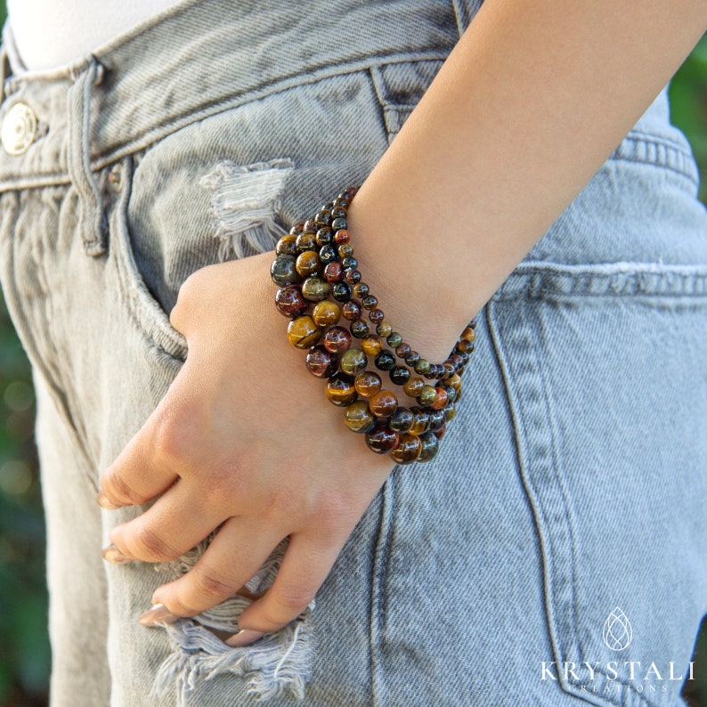 May include: A stack of six bracelets made of tiger's eye beads in shades of brown, gold, and black. The bracelets are worn on a person's wrist.