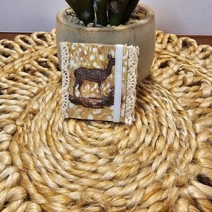 May include: A small, brown and white fabric wallet with a deer illustration and white lace trim. The wallet is sitting on a woven, tan, circular mat.