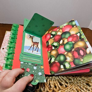 May include: A handmade Christmas scrapbook with a green cover and a red deer illustration. The scrapbook is open to reveal pages with a variety of Christmas-themed patterns, including ornaments, snowflakes, and lights.