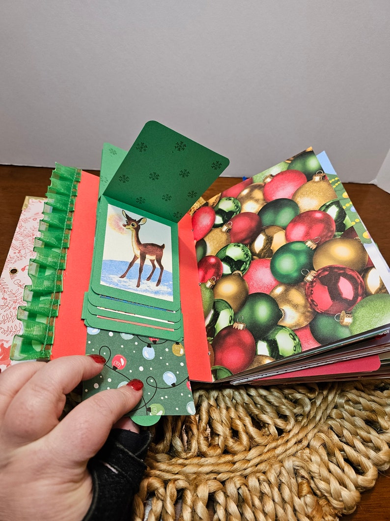 May include: A handmade Christmas scrapbook with a green cover and a red deer illustration. The scrapbook is open to reveal pages with a variety of Christmas-themed patterns, including ornaments, snowflakes, and lights.