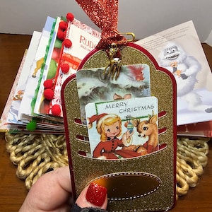 May include: A gold glitter Christmas card holder with a red ribbon and a gold reindeer charm. The card holder has a pocket for a card and a small gold tag. The card holder is decorated with a vintage Christmas card featuring a girl and a reindeer. The card reads "Merry Christmas".
