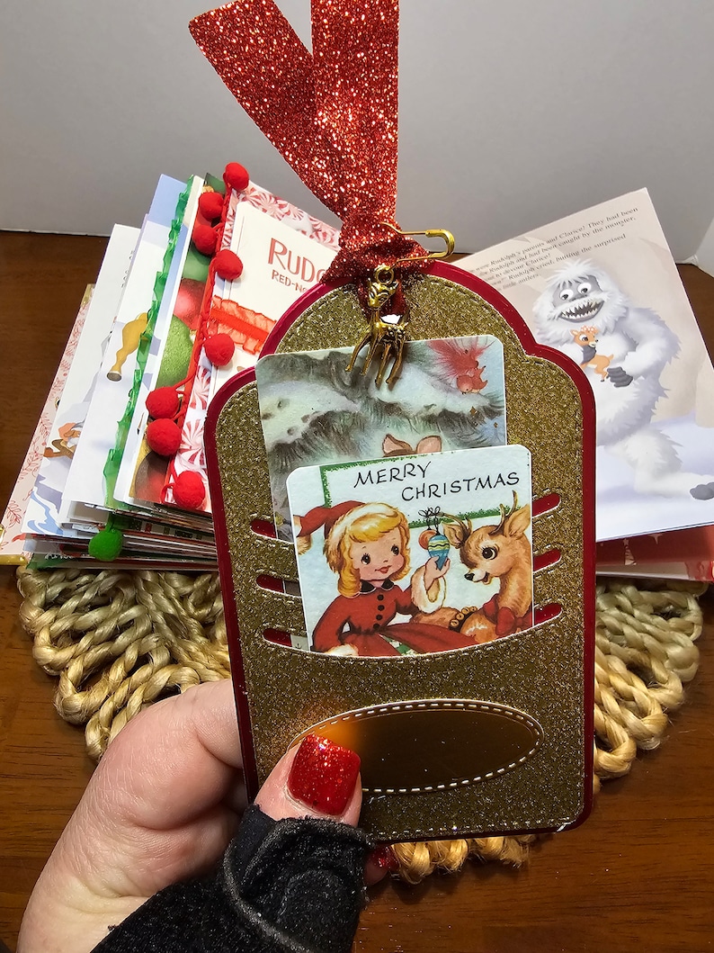 May include: A gold glitter Christmas card holder with a red ribbon and a gold reindeer charm. The card holder has a pocket for a card and a small gold tag. The card holder is decorated with a vintage Christmas card featuring a girl and a reindeer. The card reads "Merry Christmas".