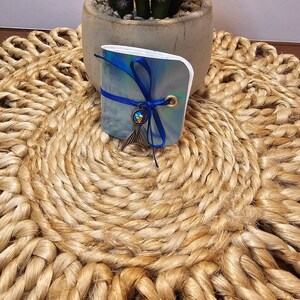 May include: A small, iridescent blue notebook tied with a dark blue ribbon and a mermaid tail charm. The notebook is resting on a woven, circular placemat. A small potted plant is in the background.
