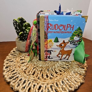 May include: A vintage copy of the children's book "Rudolph the Red-Nosed Reindeer" by the Golden Book. The book is decorated with a green ribbon, red and green plaid fabric, and gold glitter. A small cactus plant is in a white pot in the background.