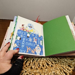 May include: A blue and white patterned scrapbook with a green cover. The scrapbook is open to reveal a page with a picture of a snowman and other Christmas characters. The text on the page reads "Clarice, Hermes, Rudolph, Sam".