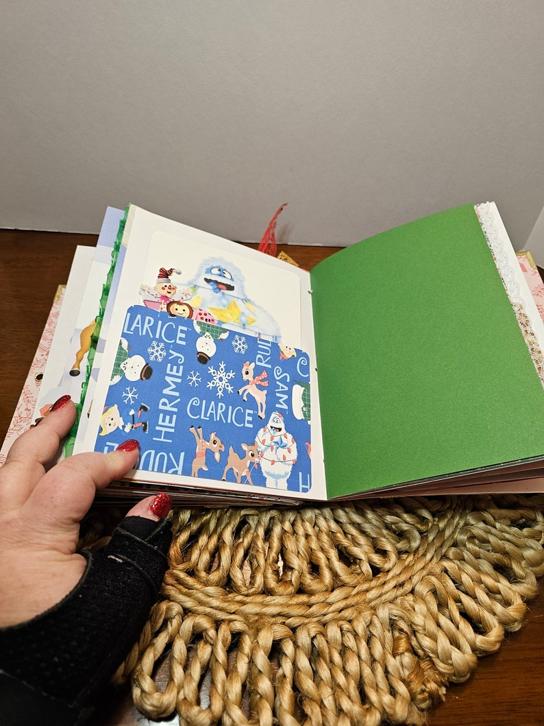 May include: A blue and white patterned scrapbook with a green cover. The scrapbook is open to reveal a page with a picture of a snowman and other Christmas characters. The text on the page reads "Clarice, Hermes, Rudolph, Sam".