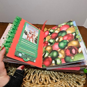 May include: An open scrapbook with a red cover and green pages. The pages are decorated with Christmas ornaments, a Rudolph the Red-Nosed Reindeer illustration, and green and red paper.