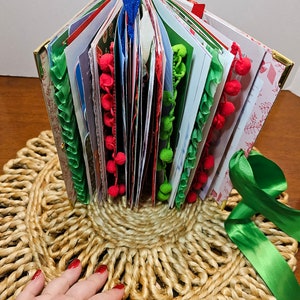 May include: A handmade Christmas journal with a green, red, and white ribbon, decorated with pom-poms and lace. The journal is open and standing upright on a woven jute mat.