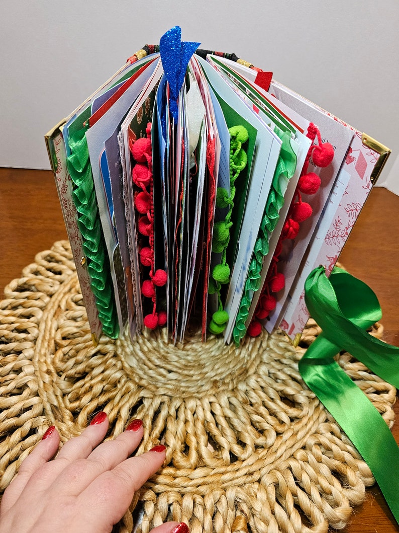 May include: A handmade Christmas journal with a green, red, and white ribbon, decorated with pom-poms and lace. The journal is open and standing upright on a woven jute mat.
