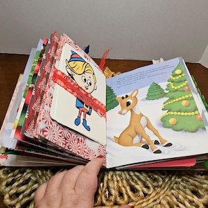 May include: An open book with a red ribbon and a Christmas theme. The book features illustrations of an elf and a reindeer. The text on the page reads: "The pair decided to be friends and to be merry together. They had wonderful games that they played in the snow, making May Belle's reindeer, Marigold, and the monster the roaring of the Arctic Sea."