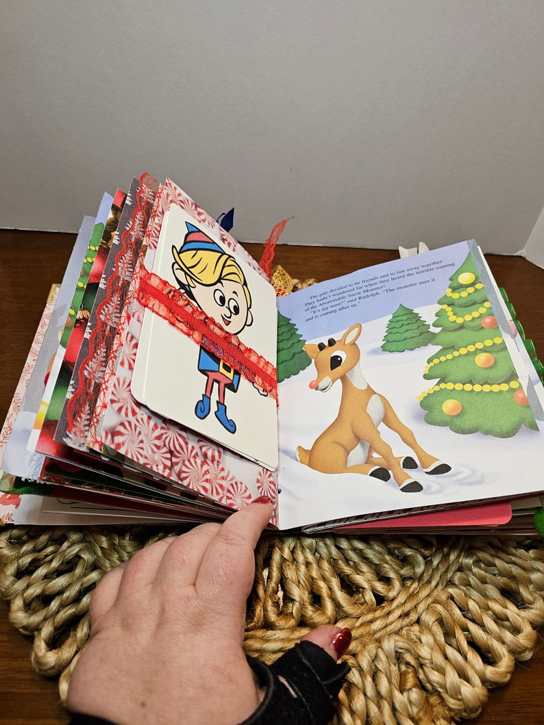 May include: An open book with a red ribbon and a Christmas theme. The book features illustrations of an elf and a reindeer. The text on the page reads: "The pair decided to be friends and to be merry together. They had wonderful games that they played in the snow, making May Belle's reindeer, Marigold, and the monster the roaring of the Arctic Sea."