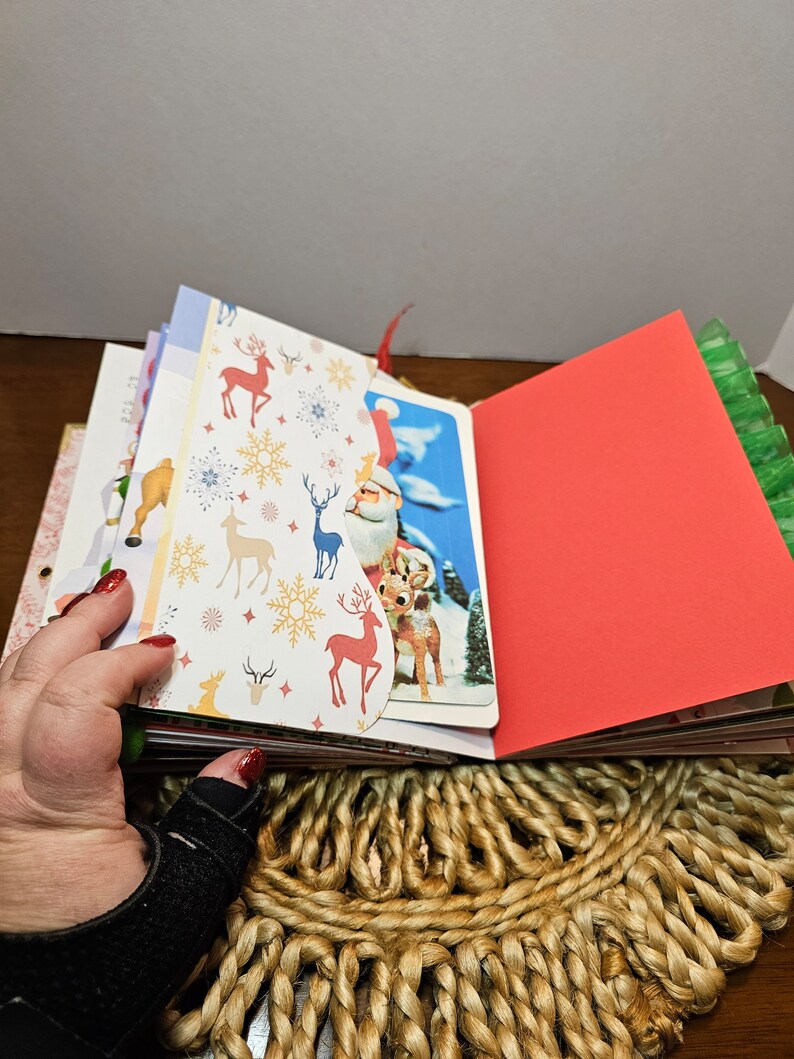 May include: A Christmas themed scrapbook with a red cover and pages decorated with reindeer and snowflakes. The scrapbook is open to reveal a page with a vintage Christmas card featuring Santa Claus and reindeer.