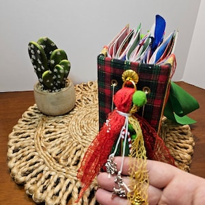 May include: A red and green plaid fabric covered journal with a red ribbon, white lace, and green and gold embellishments. The journal is decorated with a small, white, and red pom-pom.